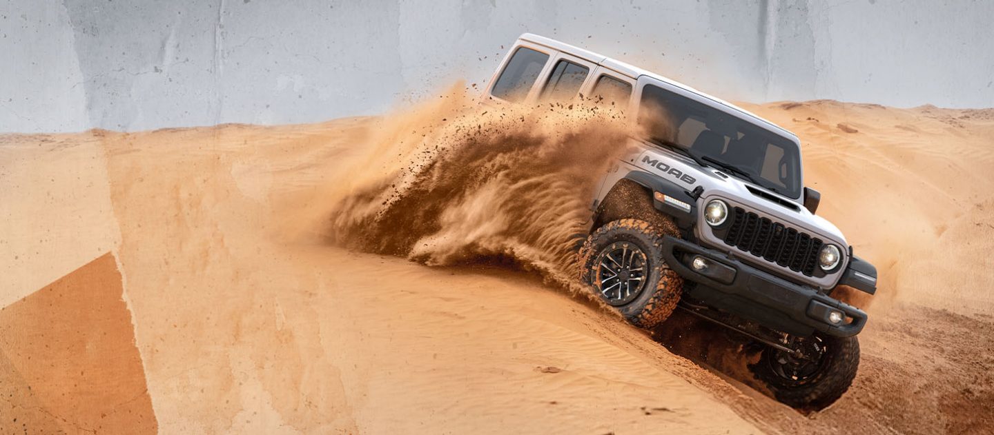 A white 2026 Jeep Wrangler Moab 392 descending a dune, kicking a cloud of sand up to its windows as it goes.