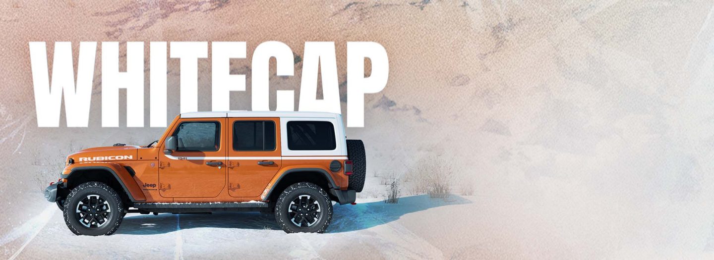 A driver-side profile of an orange 2026 Jeep Wrangler Rubicon Whitecap parked on a snow-covered clearing, with mountains in the background. Whitecap.